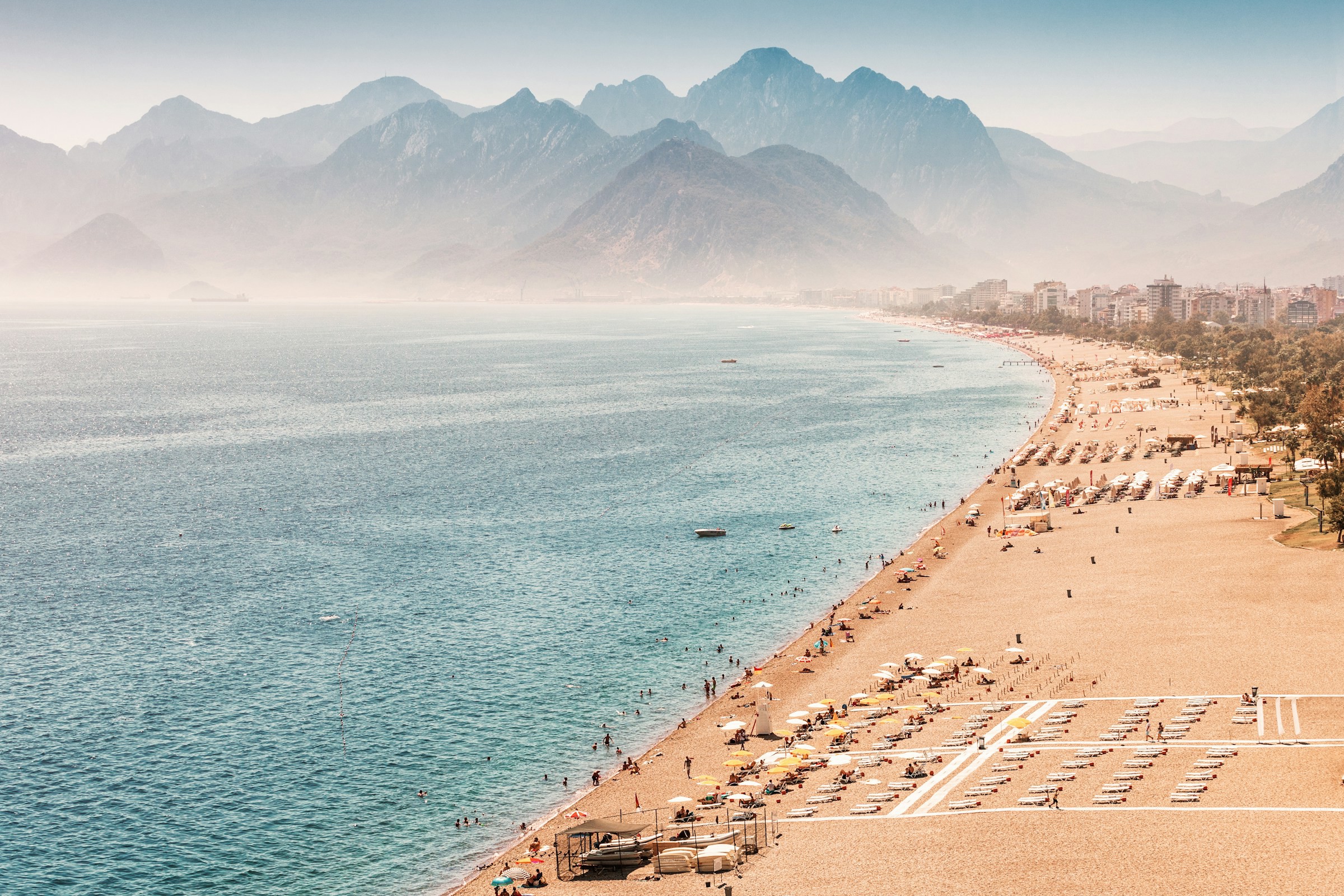 Vy över stor strand längs blått hav ut i horisonten med en stad och berg i Antalya, Turkiet