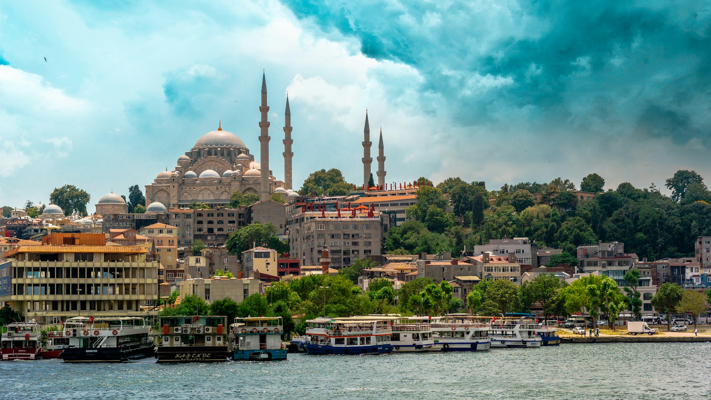 Panoramautsikt över den historiska Suleymaniye-moskén i Istanbul, med båtar förankrade längs kajen och dramatisk himmel ovanför.