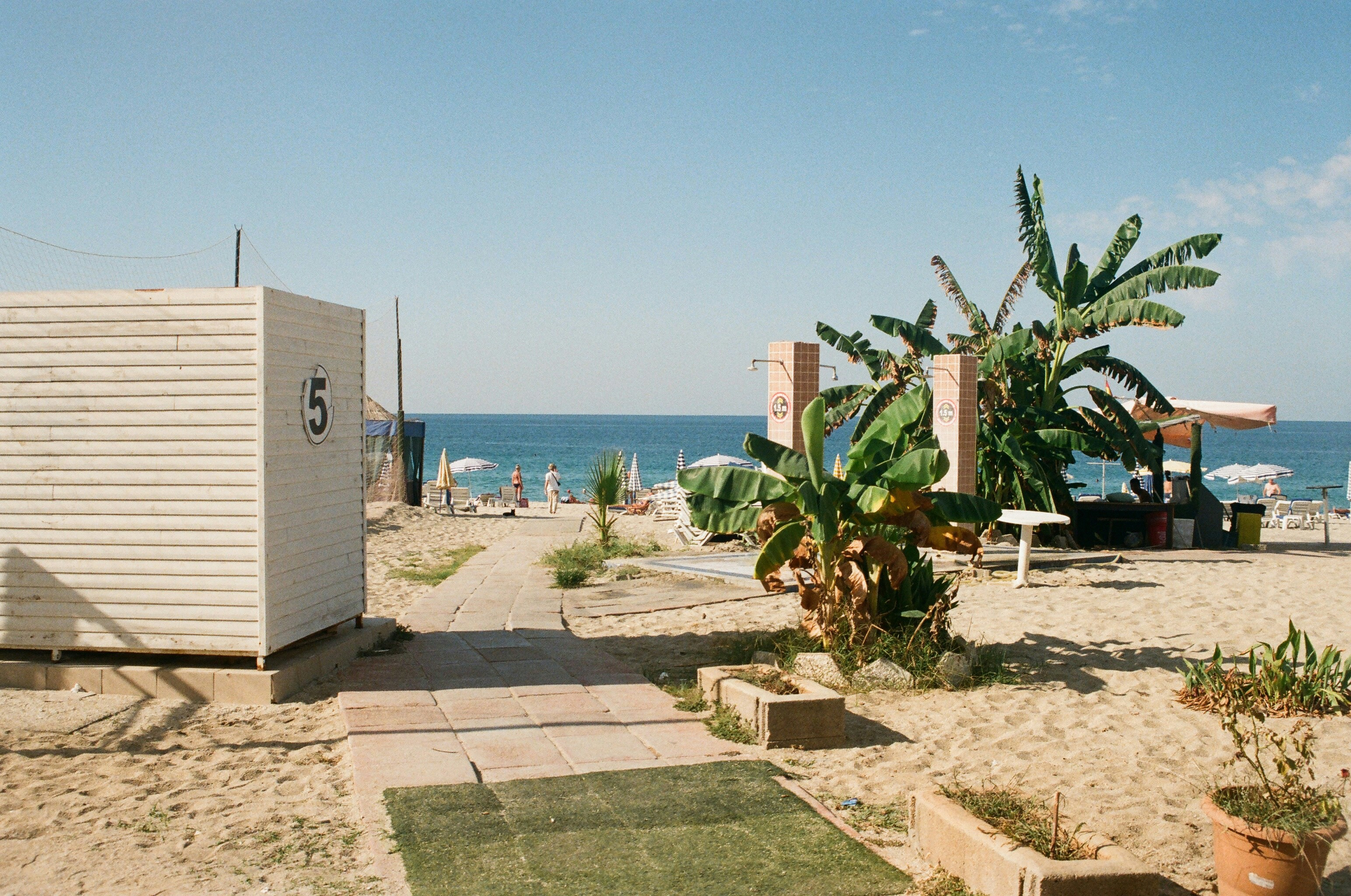 Sandig strand med badhytter, palmer och besökare vid havet på en solig dag i Alanya.