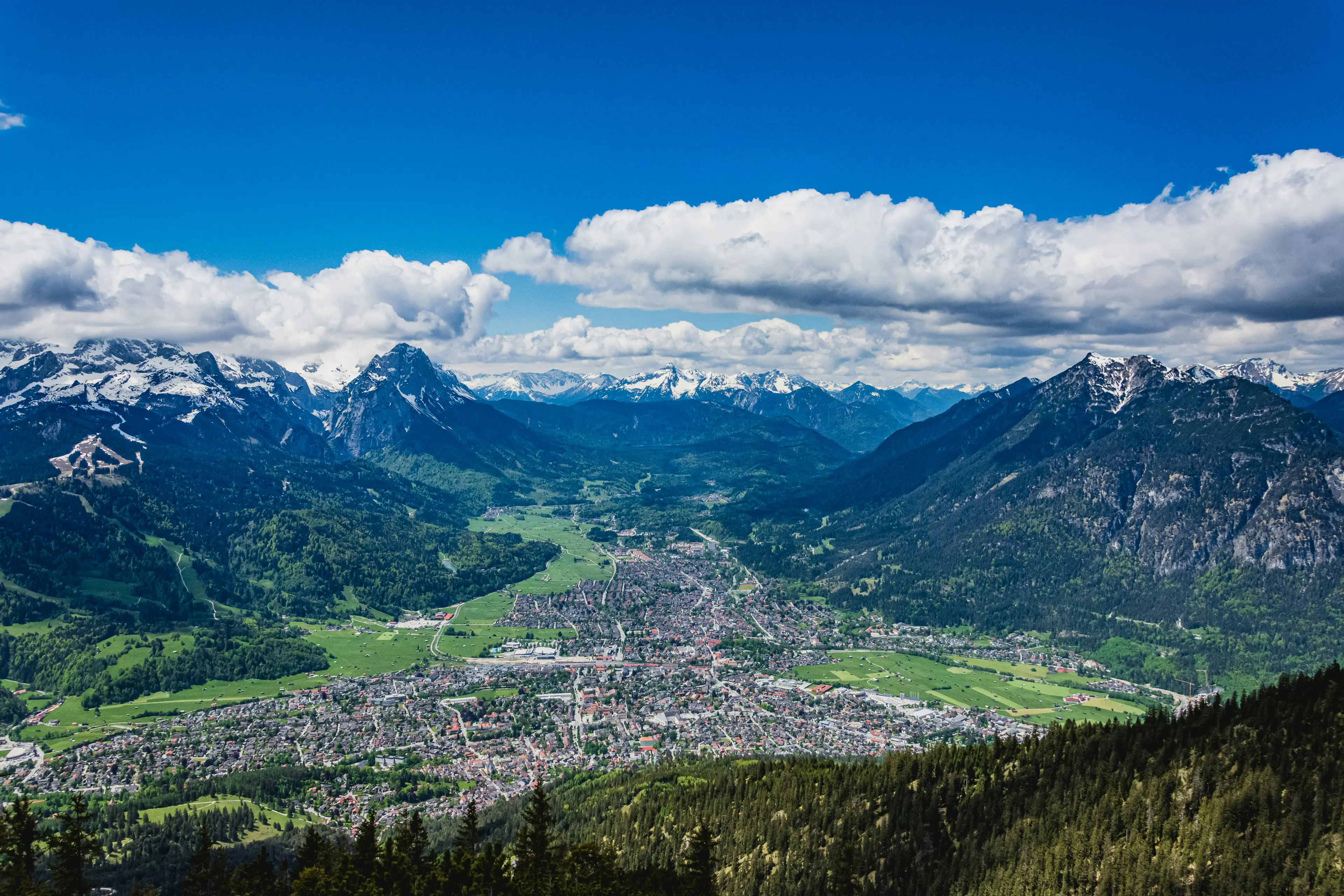 Resa till Garmisch - Panorama vy över stad mellan mellan skog och mäktiga snötäckta berg med blå himmel i bakgrunden