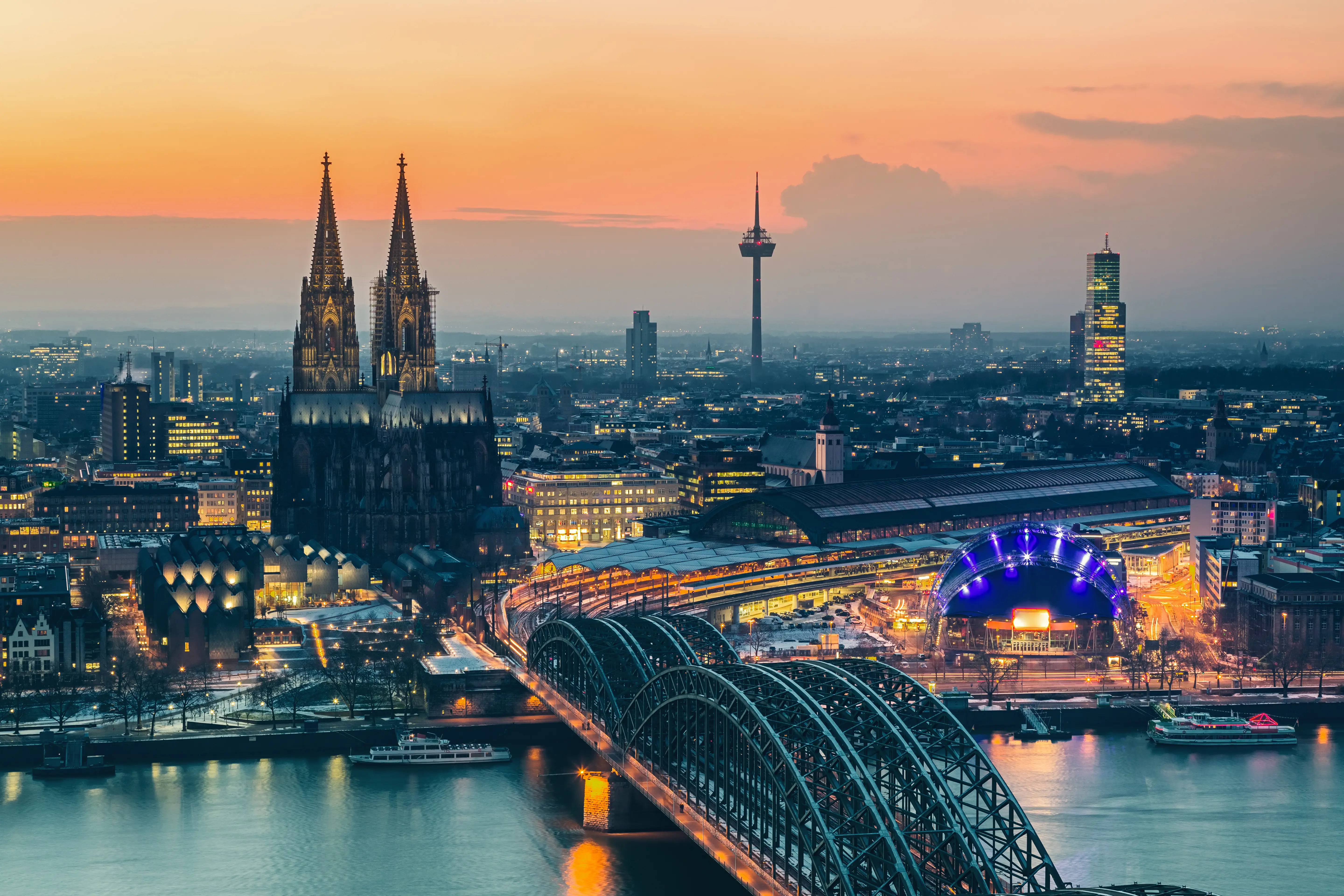 Resa till Tyskland - Panoramavy över Köln vid skymningen med Kölnerdomen, Hohenzollernbron och Rheinfloden i förgrunden, samt belysta stadsbyggnader och en dramatiskt himmel i bakgrunden.