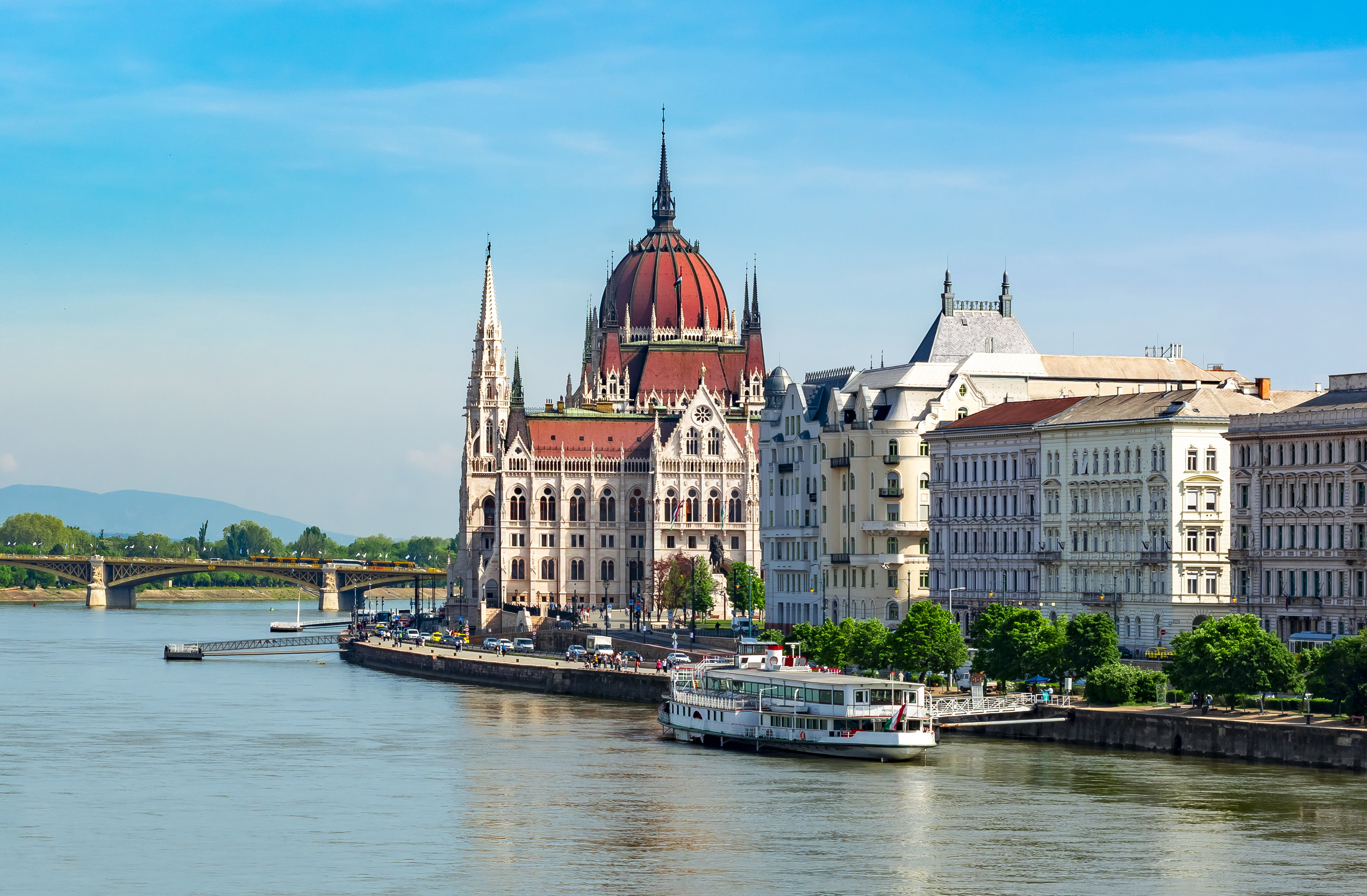 Utsikt över det ikoniska Ungerska parlamentet i Budapest vid floden Donau med en turistbåt och Margitbron i bakgrunden på en klar dag.