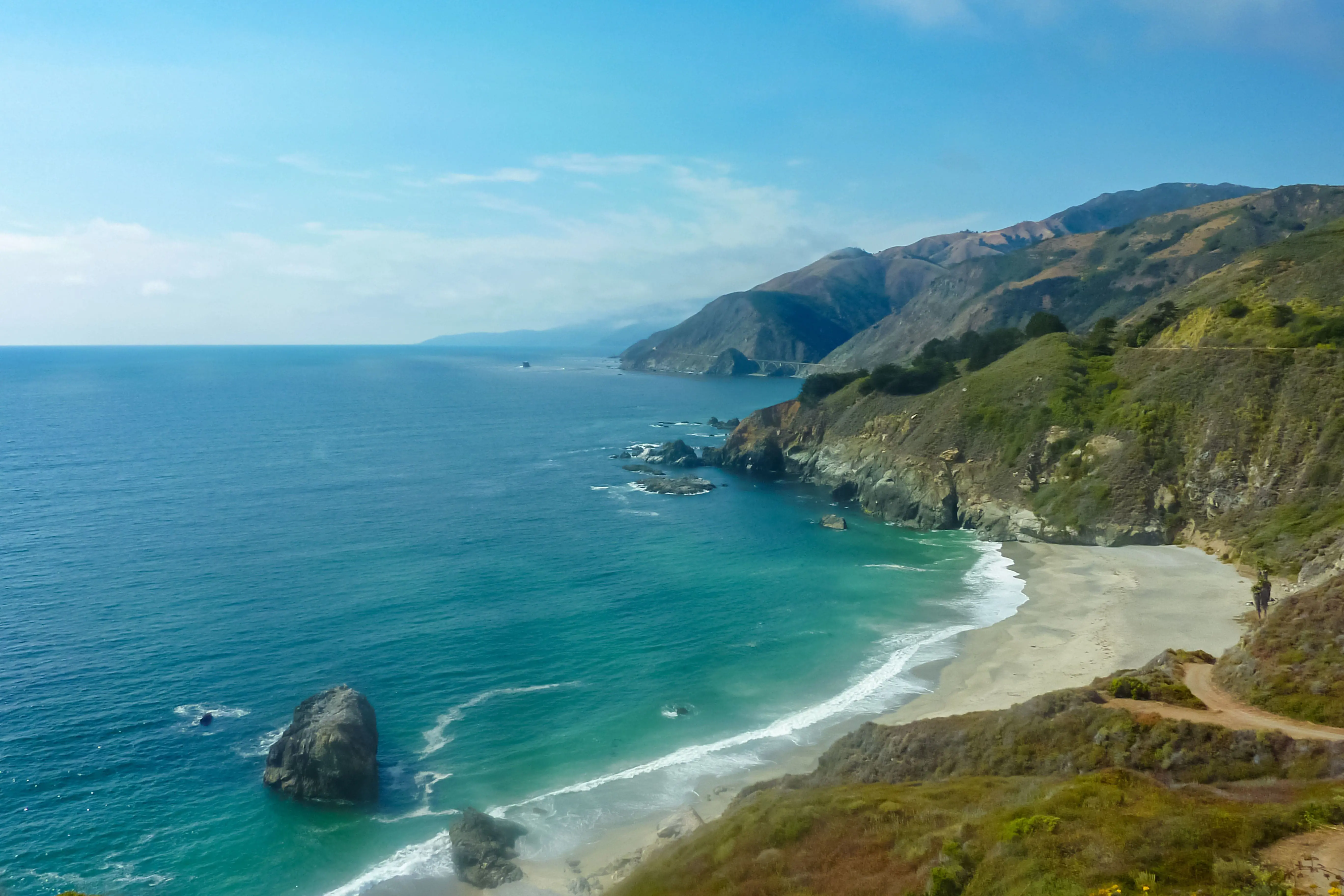 Scenic view of Big Sur coastline featuring rugged cliffs, turquoise ocean waves, and a sandy beach under a clear sky in California.