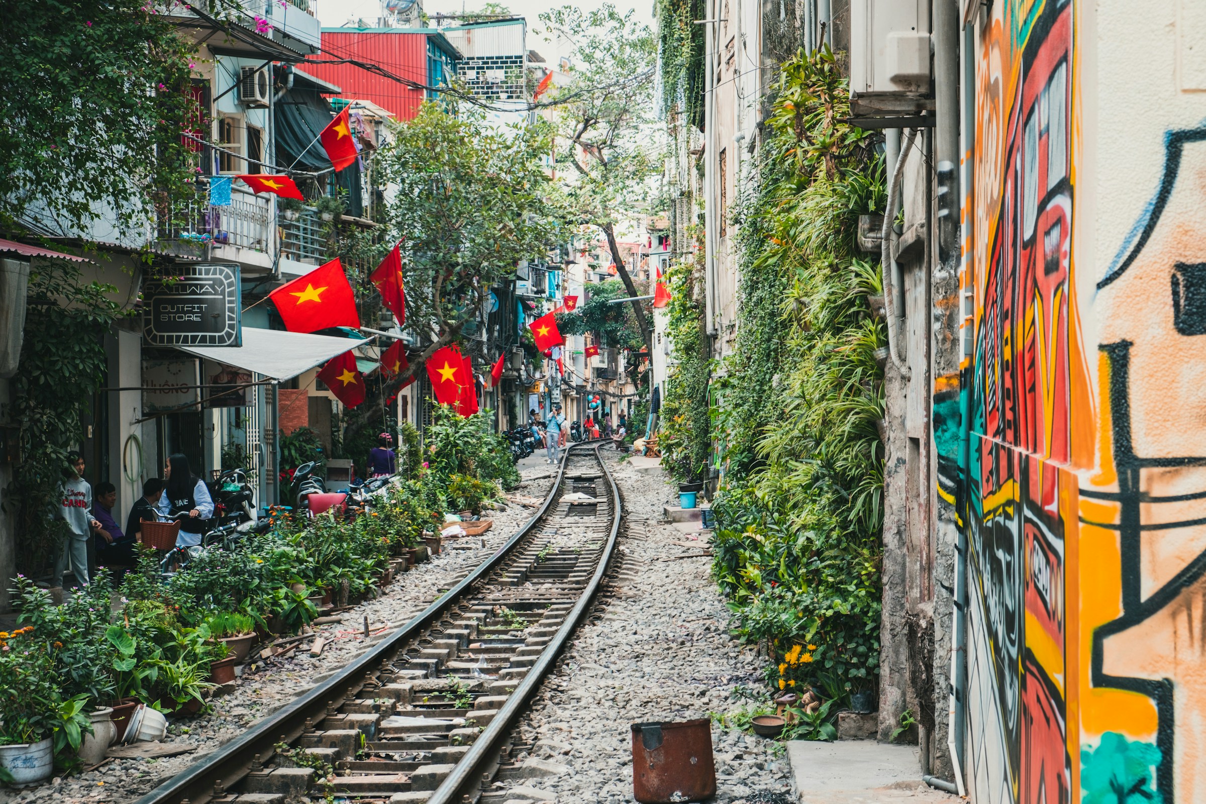 Gatumiljö med järnvägsspår genom bostadsområde med grönska och graffiti i Hanoi, dekorerad med Vietnamesiska flaggor.