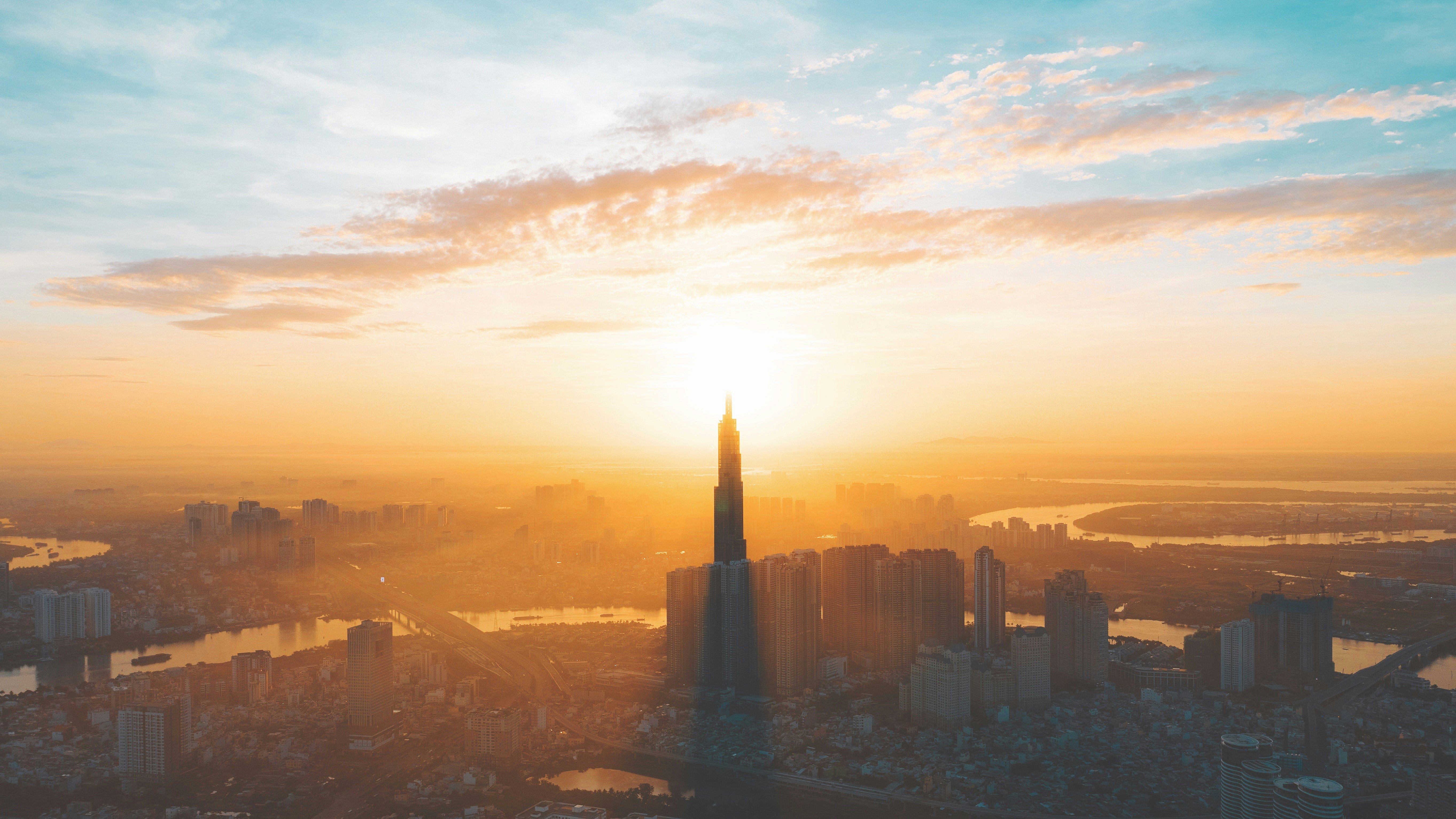 Travel to Ho Chi Minh City - Sunrise over the city skyline with towering skyscrapers and a river reflecting the morning light, with a clear sky and soft clouds in the background.