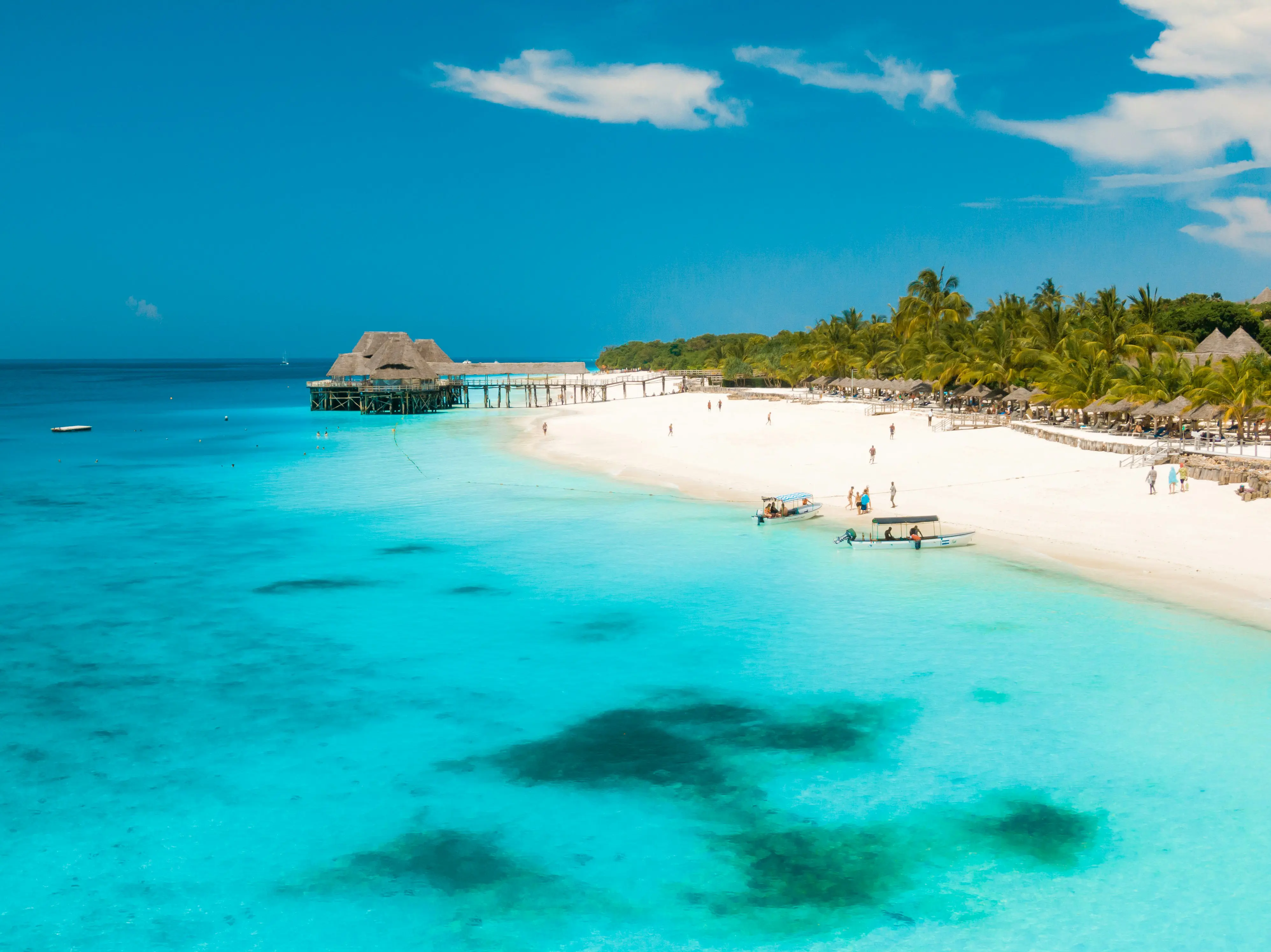 Turkosblått hav möter en vit strand med lite båtar och folk och palmer i bakgrunden på Zanzibar