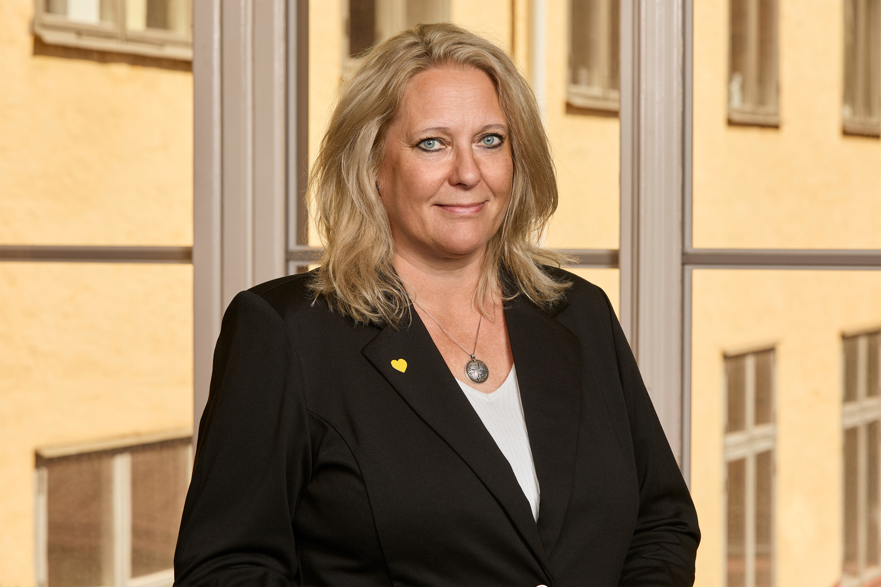 Kati Andersson - Blonde woman in a black blazer in a personal photo with a yellow building in the background