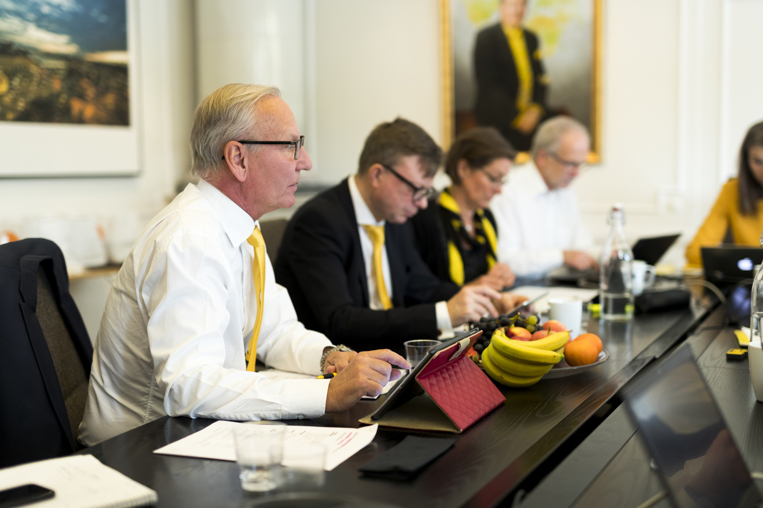 Affärsmöte med fokus på en man i förgrunden som deltar i en diskussion, med kollegor i bakgrunden vid ett konferensbord med tekniska enheter och fruktskål.