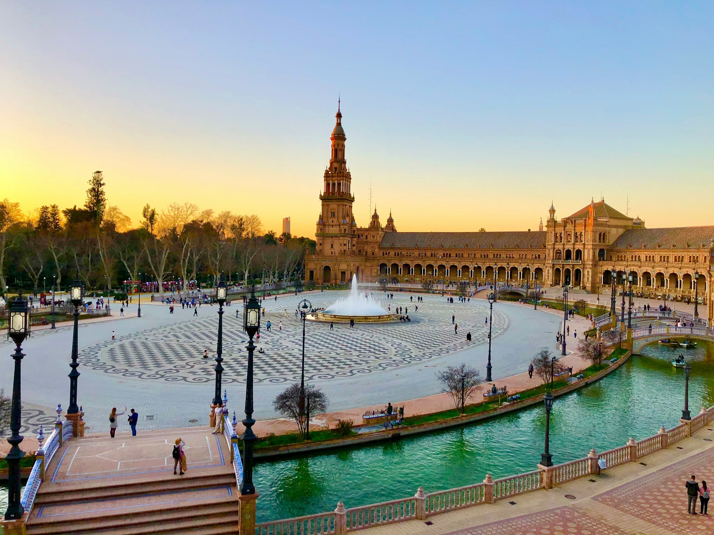 Plaza de España i Sevilla vid solnedgång med turister som promenerar och reflekterande vattenkanaler.